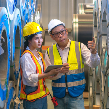 Two workers in safety gear discussing details on a tablet in front of large rolls of material in a factory setting